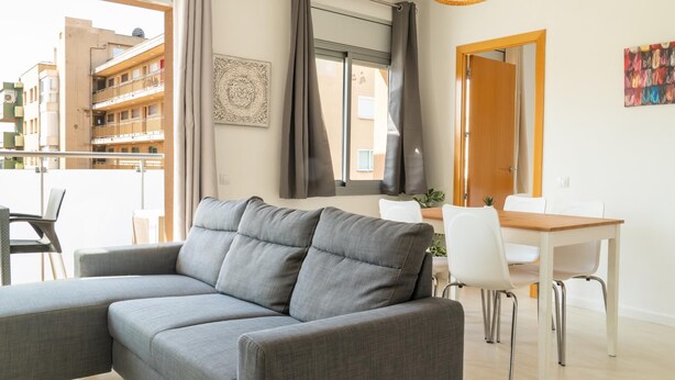 Uma sala de estar moderna com um sofá seccional cinza, uma mesa de jantar com cadeiras, um lustre e vista para edifícios.