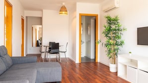 A modern living room with a grey sectional sofa, a dining area with a table and chairs, a large potted plant, and a mounted TV. - Apartaments Corall 3A (Salou)