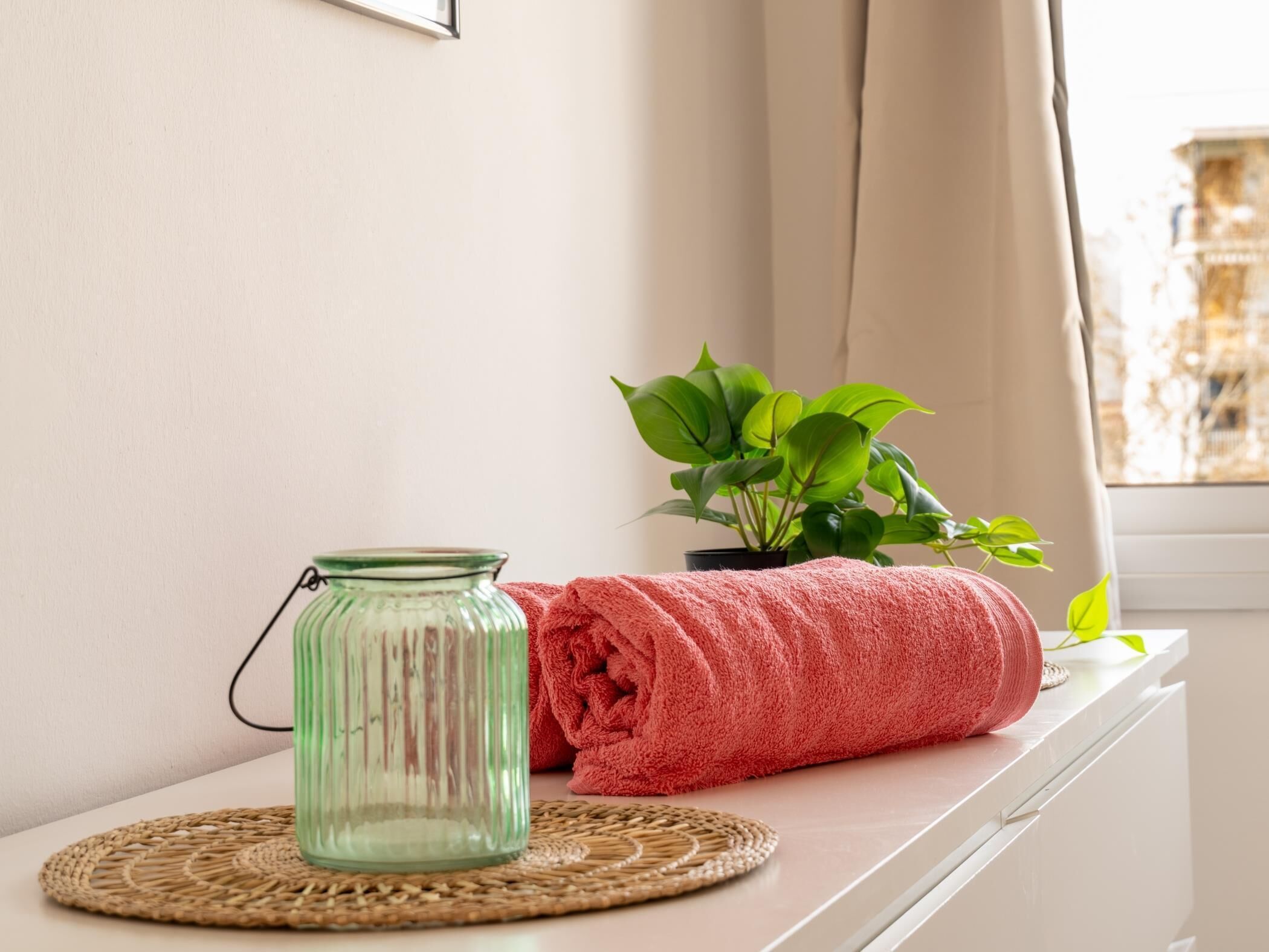 A glass vase on a woven placemat, a rolled towel, and a potted plant on a windowsill.