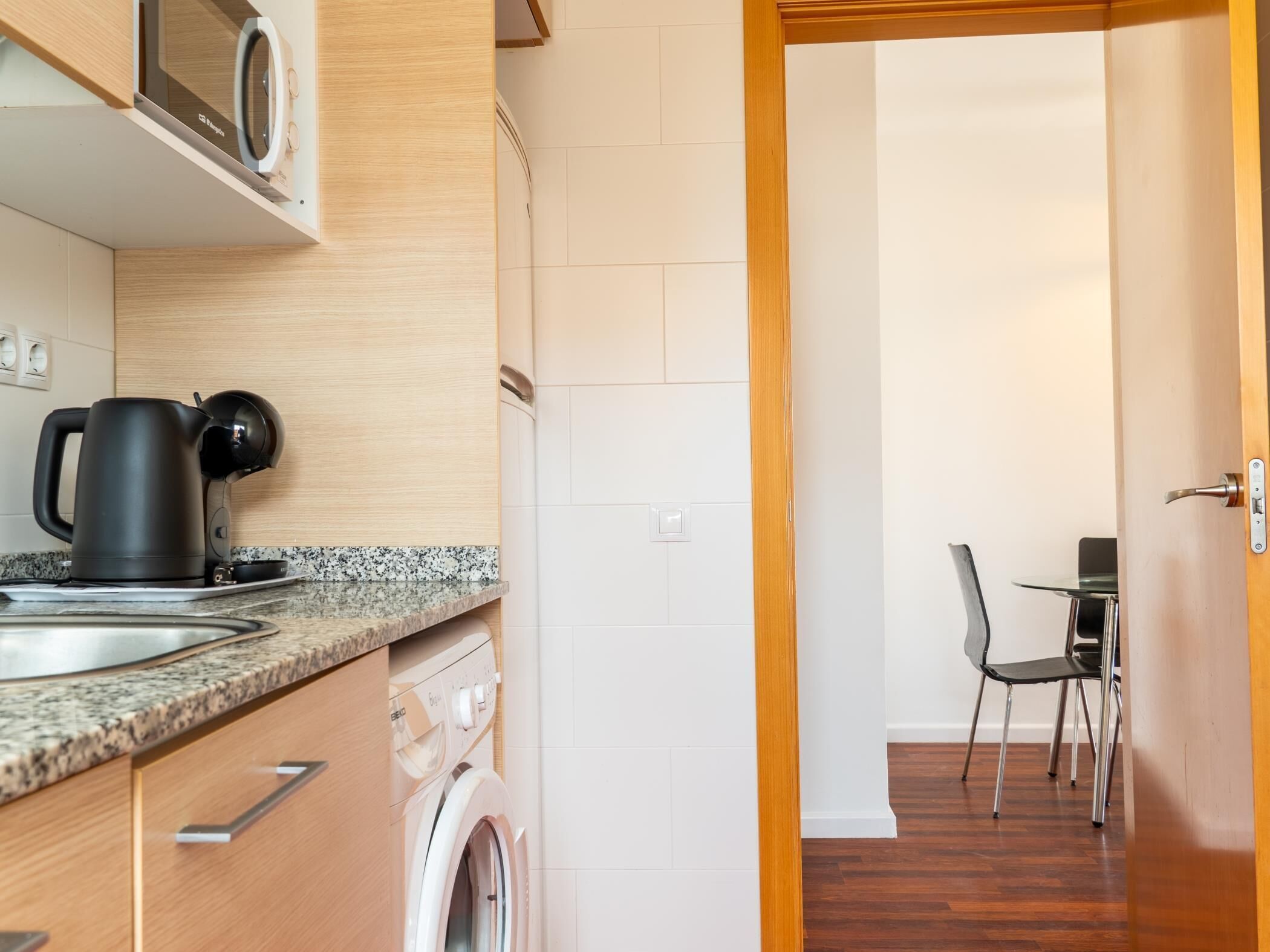A compact kitchen with a washing machine, a coffee maker, and a dining area with a chair.