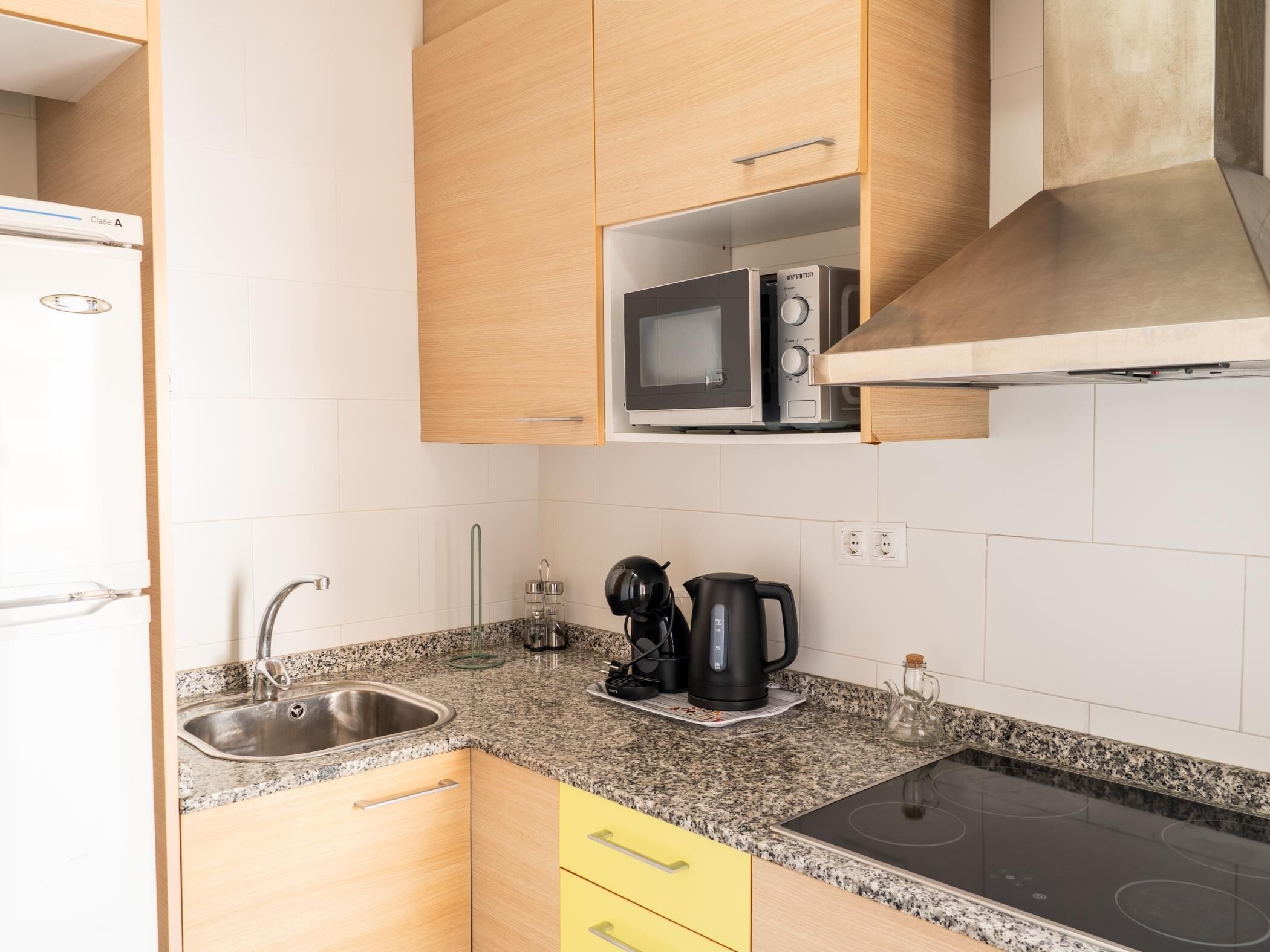Una cocina con microondas, hervidor y cafetera sobre la encimera, un lavamanos de acero inoxidable y armarios de madera.
