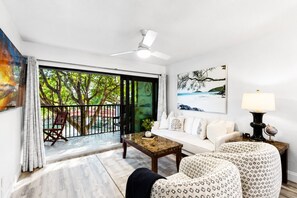 A living room with a sofa, chairs, a coffee table, a ceiling fan, and a view through a sliding glass door to an outdoor area with a tree and a red umbrella. - Park Shore Resort 2nd Floor BLDGJ*Waterviews* (Naples)
