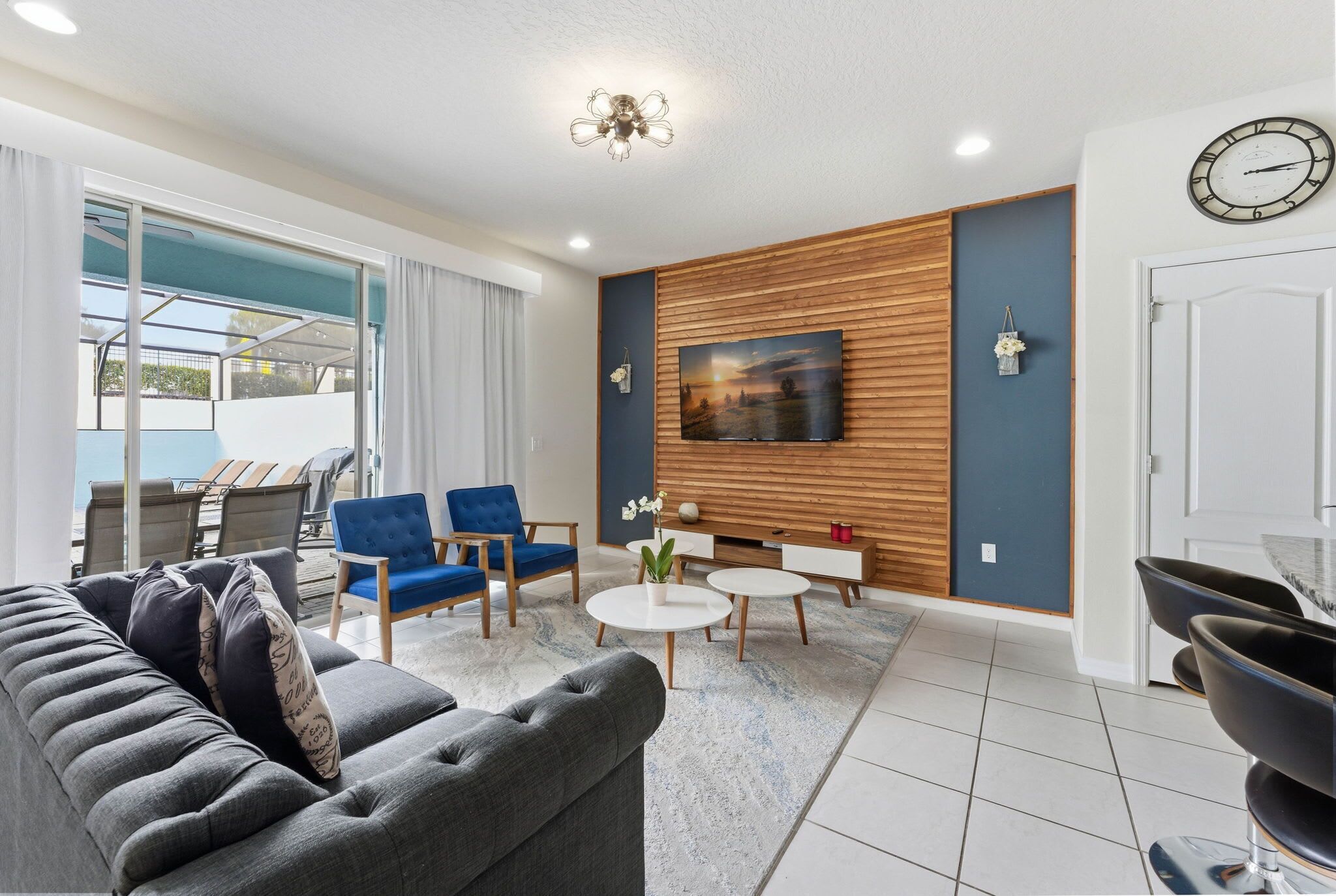 A modern living room with a grey sofa, blue armchairs, a coffee table, and a TV wall-mounted above a wooden panel.