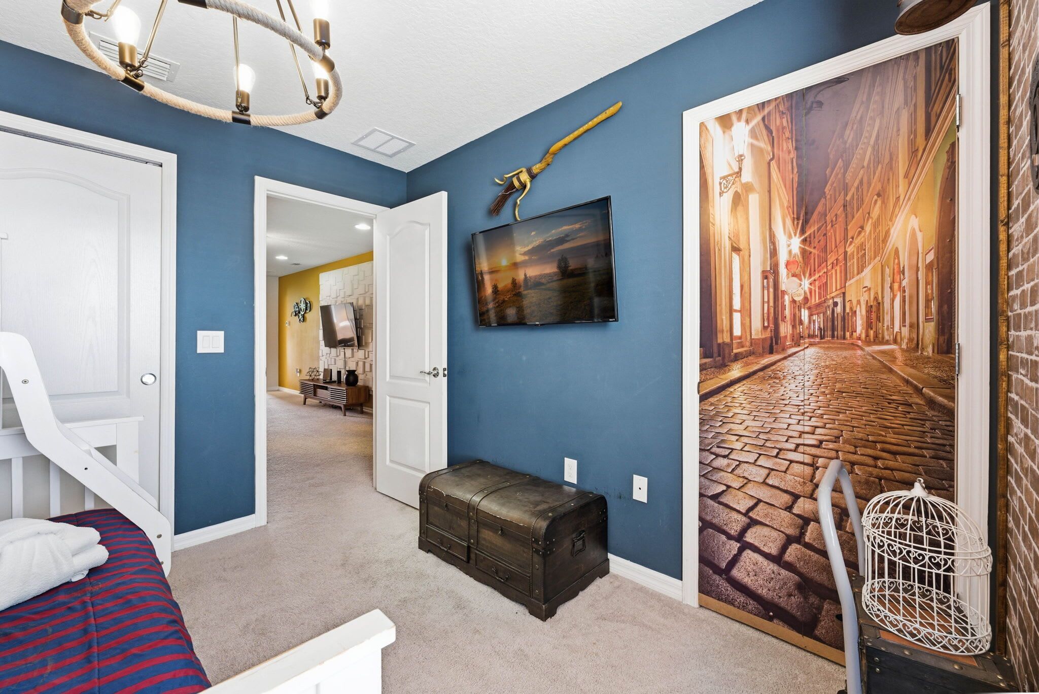 A bedroom with a bed, a chest of drawers, a suitcase, a painting, and a chandelier.