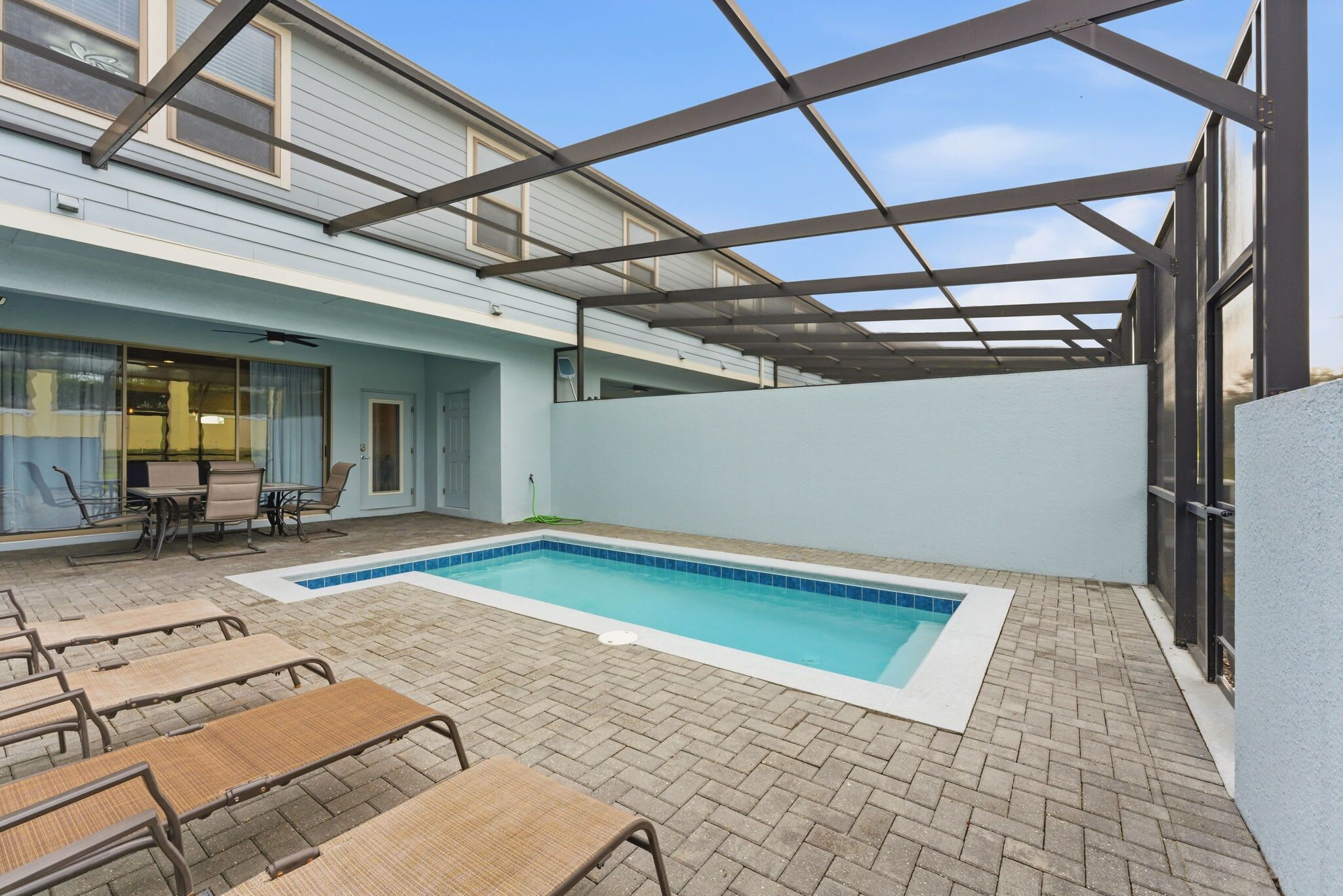 A covered outdoor swimming pool with lounge chairs and a table, surrounded by a brick patio.