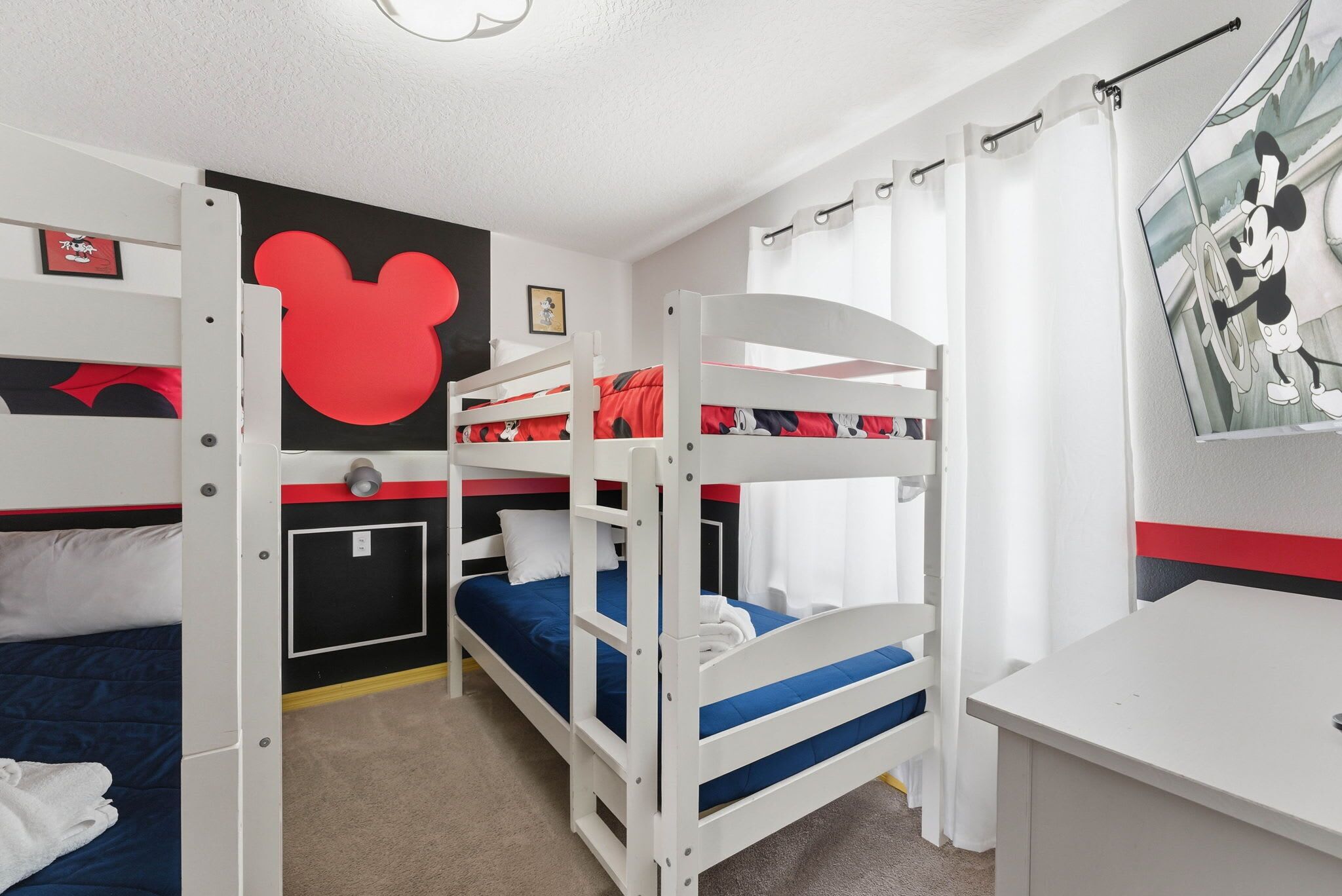 A bunk bed room with a Mickey Mouse cutout on the wall, a desk, and a window with curtains.