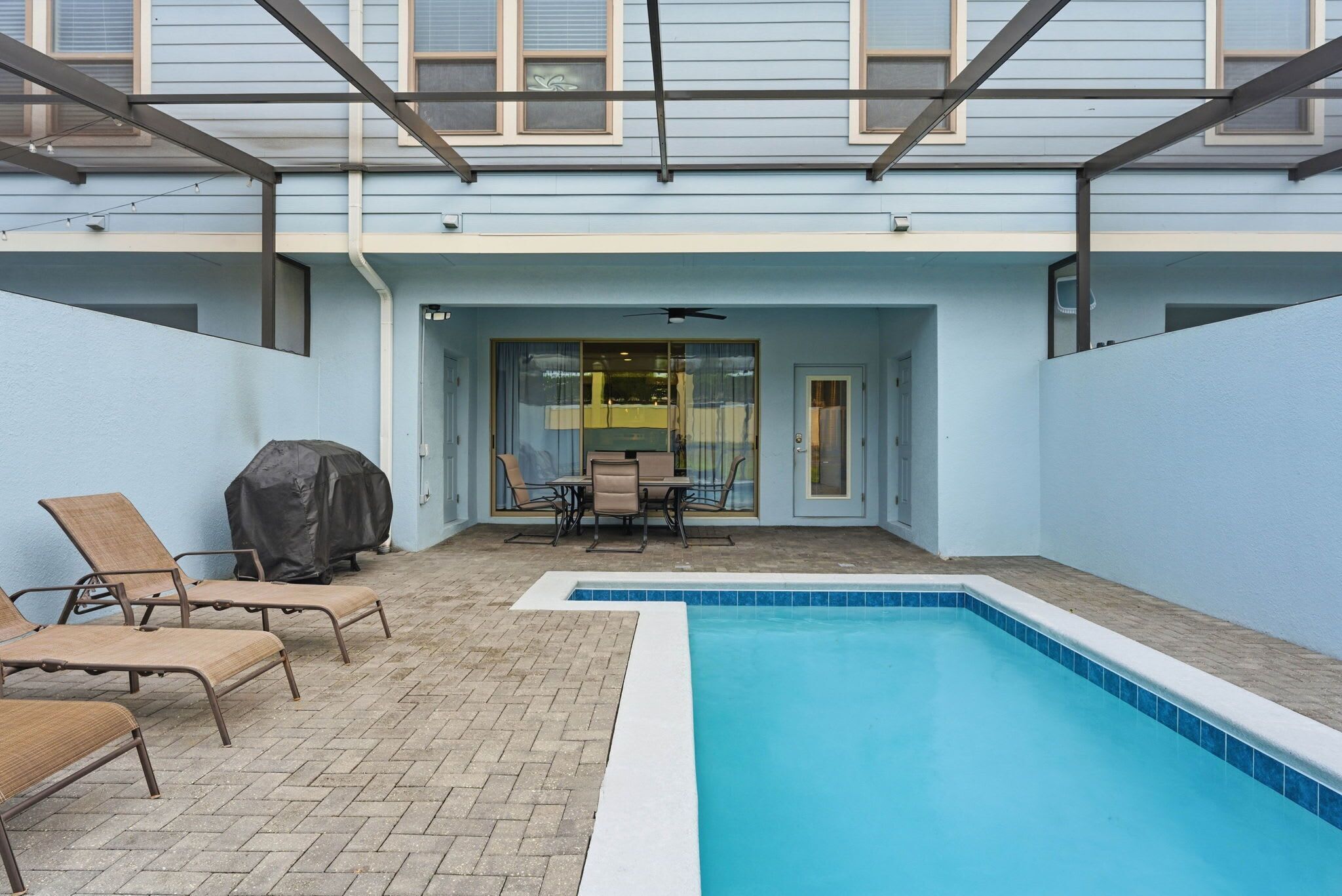 A swimming pool with lounge chairs and a covered grill area, enclosed by a building with large windows.