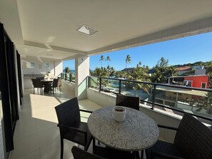 A balcony with a round table and chairs, a view of the beach and palm trees, and a kitchen area in the background. - Flat Gold Vista Mar - Praia de Tabatinga (Conde)