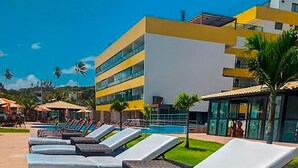A row of sun loungers with cushions, a poolside area, and a modern building in the background. - Flat Gold Vista Mar - Praia de Tabatinga (Conde)