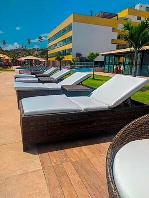 A row of sun loungers with cushions, a poolside area, and a modern building in the background. - Flat Gold Vista Mar - Praia de Tabatinga (Conde)