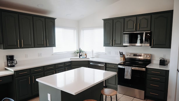 A kitchen with dark green cabinets, a white island, stainless steel appliances, and a window with blinds. - Spacious 6BR 3BA • Sleeps 16 • Across from Large Park (West Jordan)