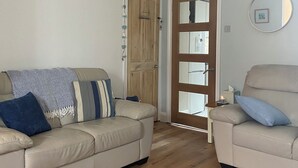A living room with a beige sofa, a smaller armchair, a wooden floor, and a door leading to another room.