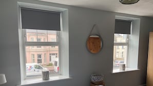 A bedroom with two windows, a bed with patterned bedding, a bedside table with a lamp, and a round mirror on the wall.