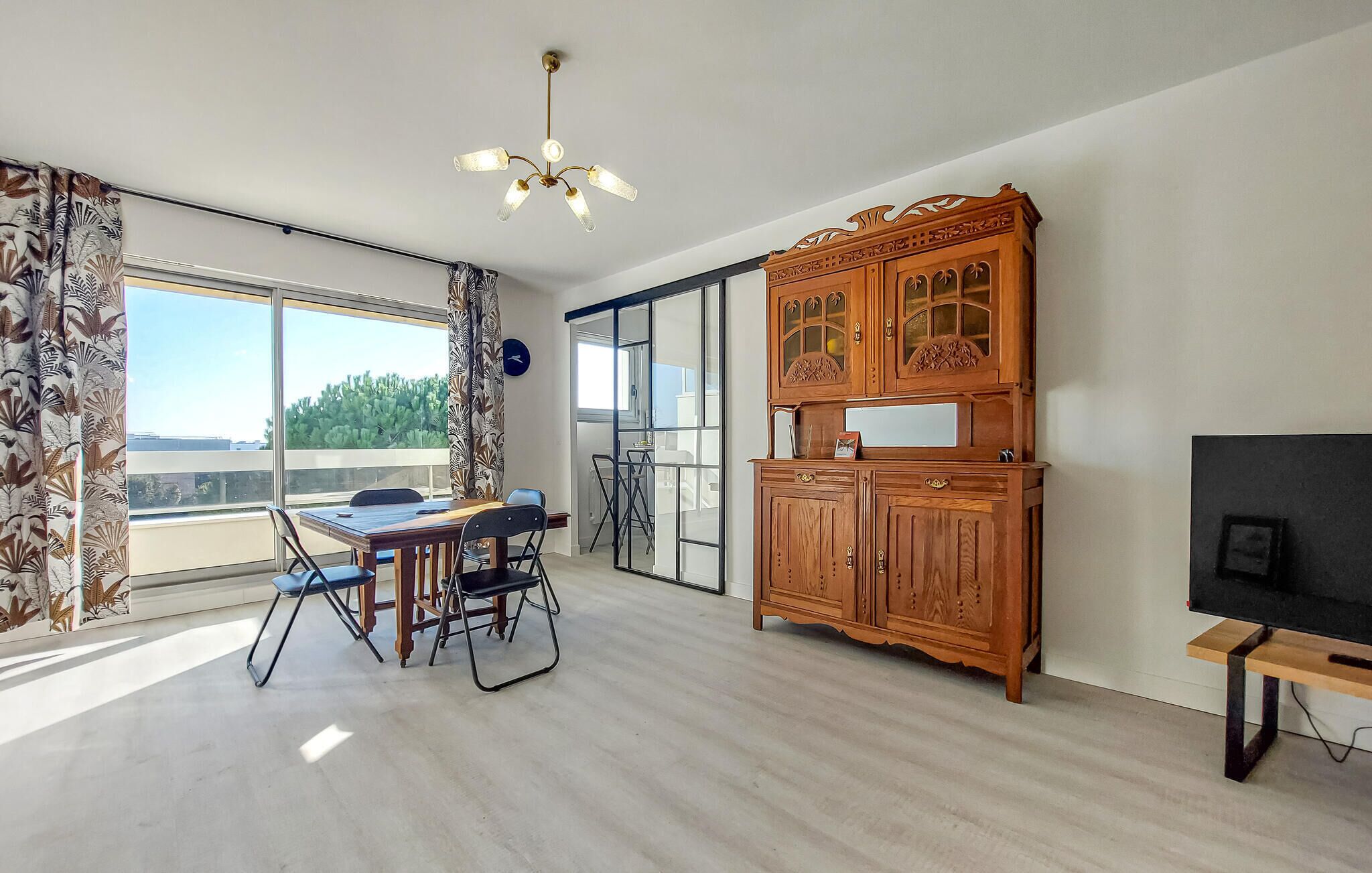 A spacious room with a dining table, chairs, a wooden cabinet, a television, and a large window with a view of greenery.