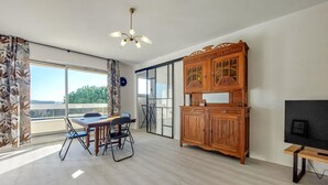 Ein geräumiges Zimmer mit Esstisch, Stühlen, einer Holzkommode, einem Fernseher und einem großen Fenster mit Blick auf Grünflächen.