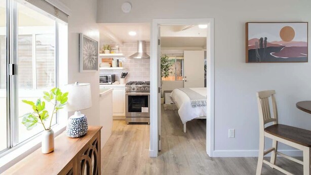Una cocina moderna con armarios blancos, un microondas y un horno. Hay una zona de comedor con una mesa y sillas de madera, y una cama al fondo.