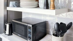 Una cocina con estantes blancos, un microondas, una cafetera y diversos utensilios de cocina.