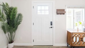 Una puerta blanca con ventanal, un cartel de bienvenida, una planta en maceta y un mueble de madera con diseño geométrico.