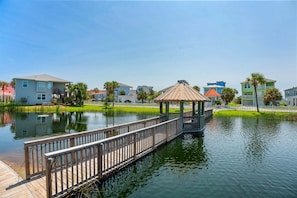 Eine Holzstege führt zu einem Pavillon über dem Wasser, umgeben von Wohnhäusern.