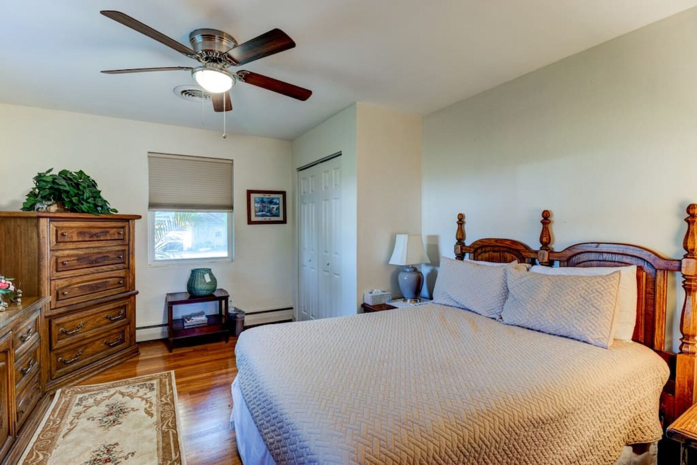 A bedroom with a large bed, wooden headboard, a ceiling fan, a nightstand with a lamp, a dresser, and a window with blinds.