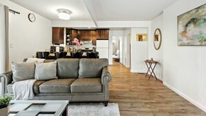 A modern living room with a grey leather sofa, wooden flooring, and a kitchen in the background. - Marbella Lane Natural Bayview Escape Near SFO (Daly City)