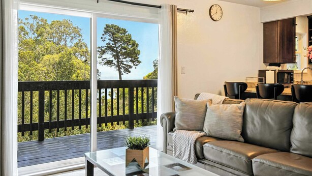 A living room with a sliding glass door to a balcony, a sofa, a coffee table, and a kitchen area with a clock on the wall. - Marbella Lane Natural Bayview Escape Near SFO (Daly City)