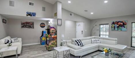 A spacious living room with a white sectional sofa, a glass coffee table, and a white couch. There are arcade games in the corner, a painting on the wall, and a door with a sign.
