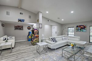 A spacious living room with a white sectional sofa, a glass coffee table, and a white couch. There are arcade games in the corner, a painting on the wall, and a door with a sign.