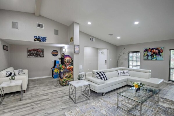 A spacious living room with a white sectional sofa, a glass coffee table, and a white couch. There are arcade games in the corner, a painting on the wall, and a door with a sign.