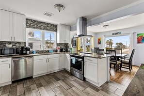 A modern kitchen with white cabinets, stainless steel appliances, a large island with seating, and a view of palm trees. - Three Oars Beach Surf Shack Pool, Oceanview, Game Room (Daytona Beach)