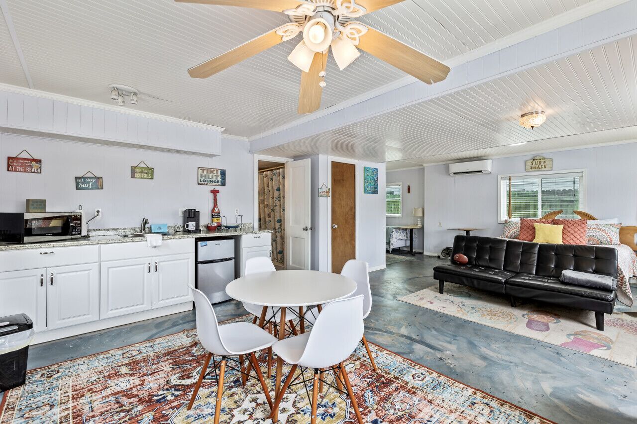 A spacious living area with a dining table, white chairs, a black leather sofa, and a ceiling fan.