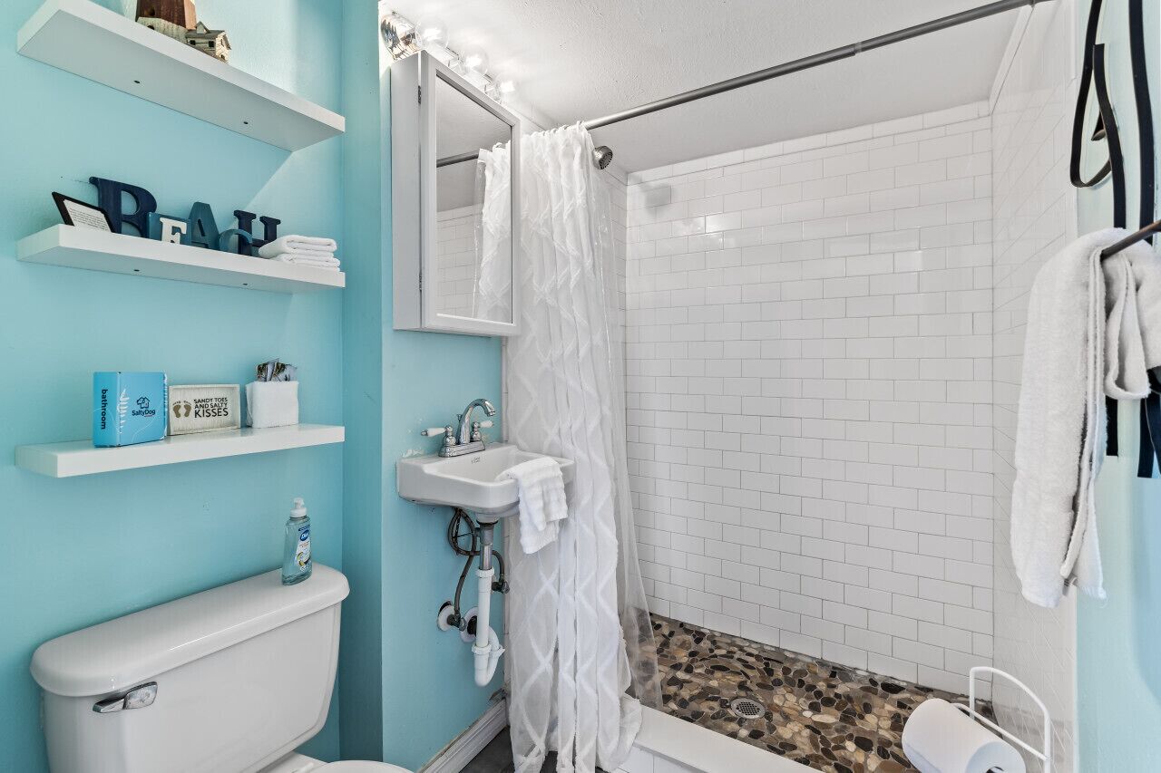 Een badkamer met toilet, wastafel en douche. Er staan planken met toiletartikelen en er hangt een spiegel.