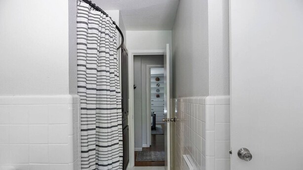 A narrow bathroom with white tiled walls, a striped shower curtain, and a visible toilet in the background. - Play & Stay Dallas Home w/ Gym + Gameroom + Wifi (Dallas)
