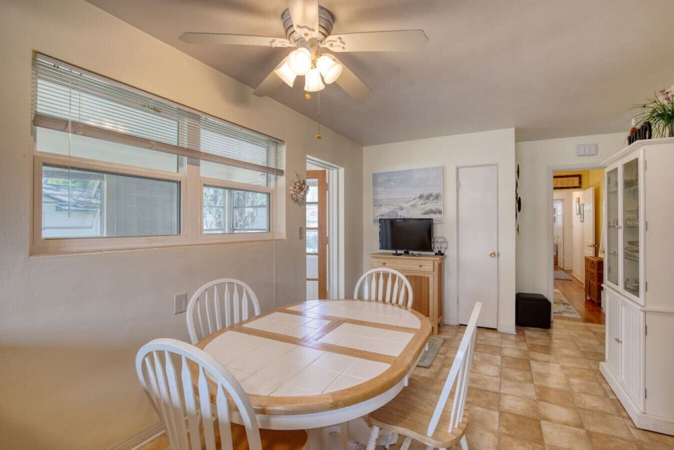 A dining area with a round table, white chairs, a ceiling fan, and a television on a cabinet.