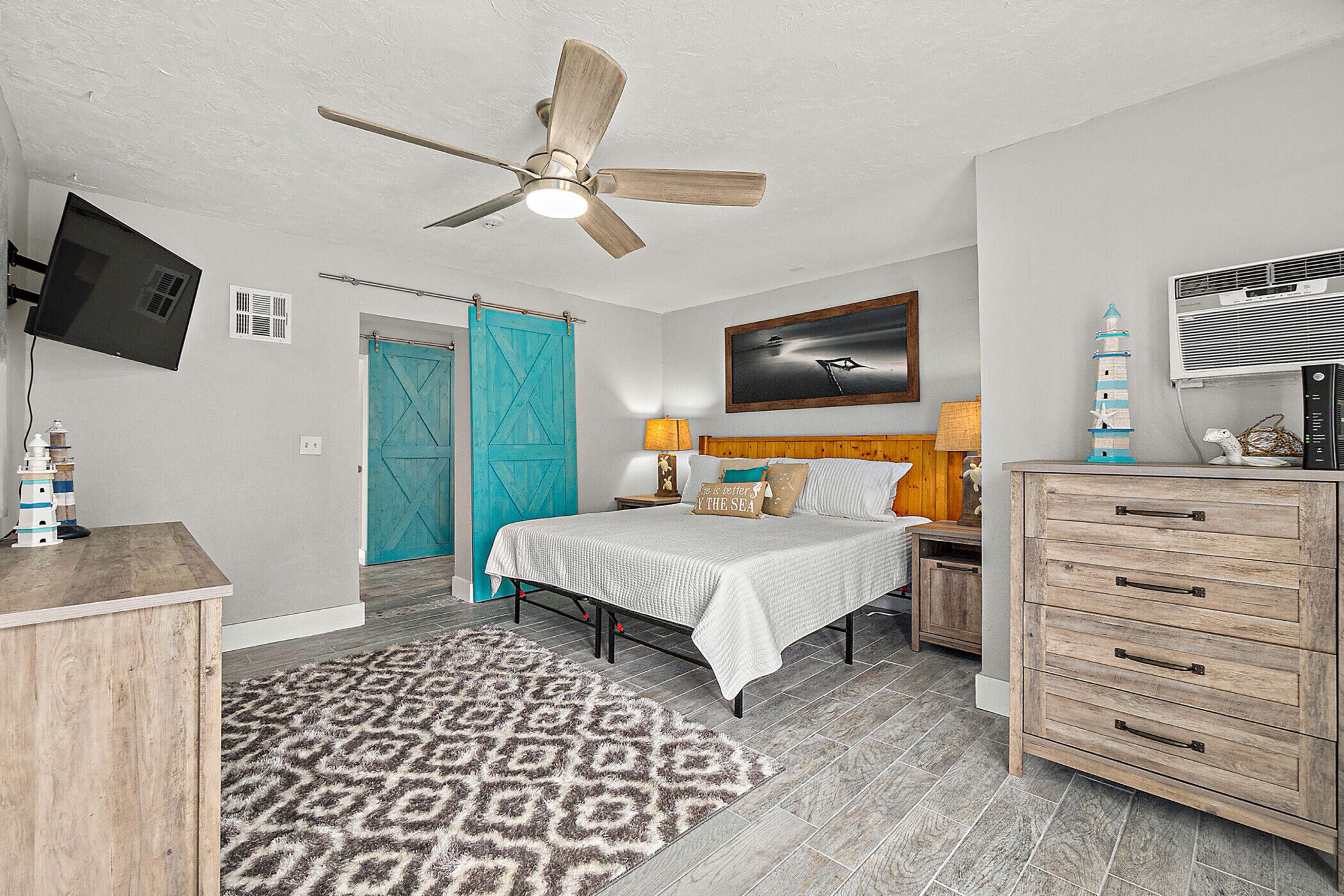 A bedroom with a bed, a ceiling fan, a TV, a dresser, a nightstand, a lamp, and a wall-mounted air conditioner.