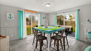 Uma área de refeições com mesa posta para seis pessoas, rodeada por cadeiras, e vista para o exterior através de portas de vidro.