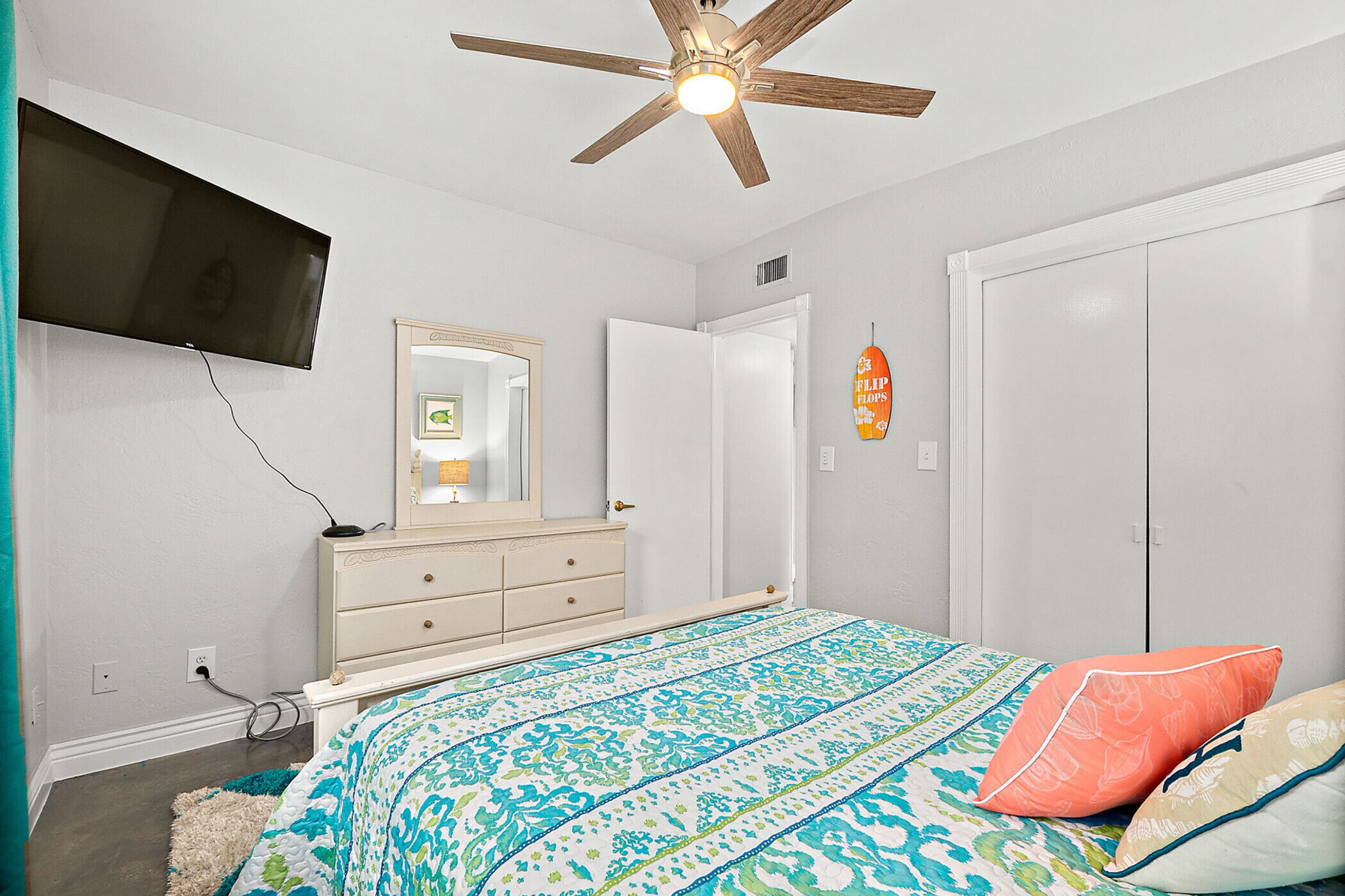 A bedroom with a bed, dresser, TV, ceiling fan, and wardrobe.