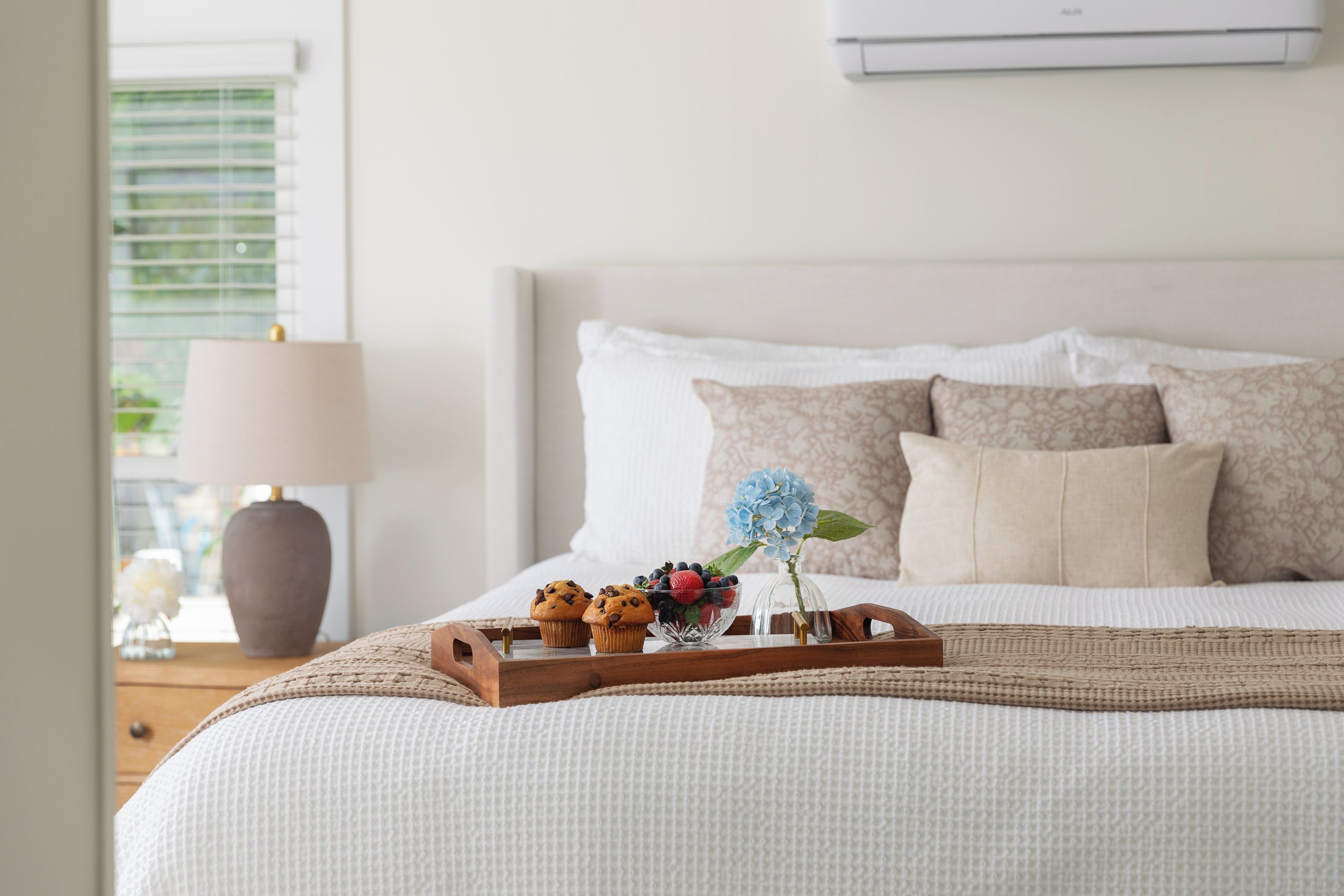 A neatly made bed with a tray of muffins and a vase of flowers, a lamp, and a window with blinds.
