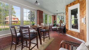 Une terrasse couverte avec une table et des chaises, un fauteuil en osier et une vue sur un bâtiment en briques.