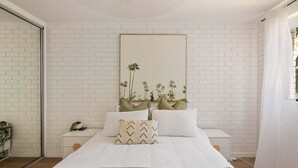 Ein Schlafzimmer mit weißer Backsteinwand, einem Bett mit weißer Bettwäsche und grünen Kissen, einem Nachttisch mit Pflanze und einem gerahmten Bild über dem Bett.
