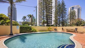 Ein Pool mit befliesster Kante, umgeben von einer Pflasterterrasse, Palmen und hohen Gebäuden im Hintergrund.