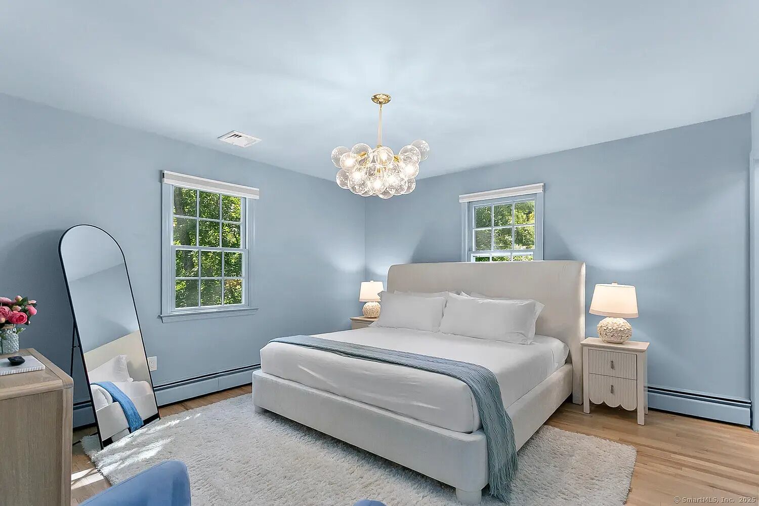 A bedroom with a large bed, two bedside tables, a chandelier, and windows with a view of greenery.