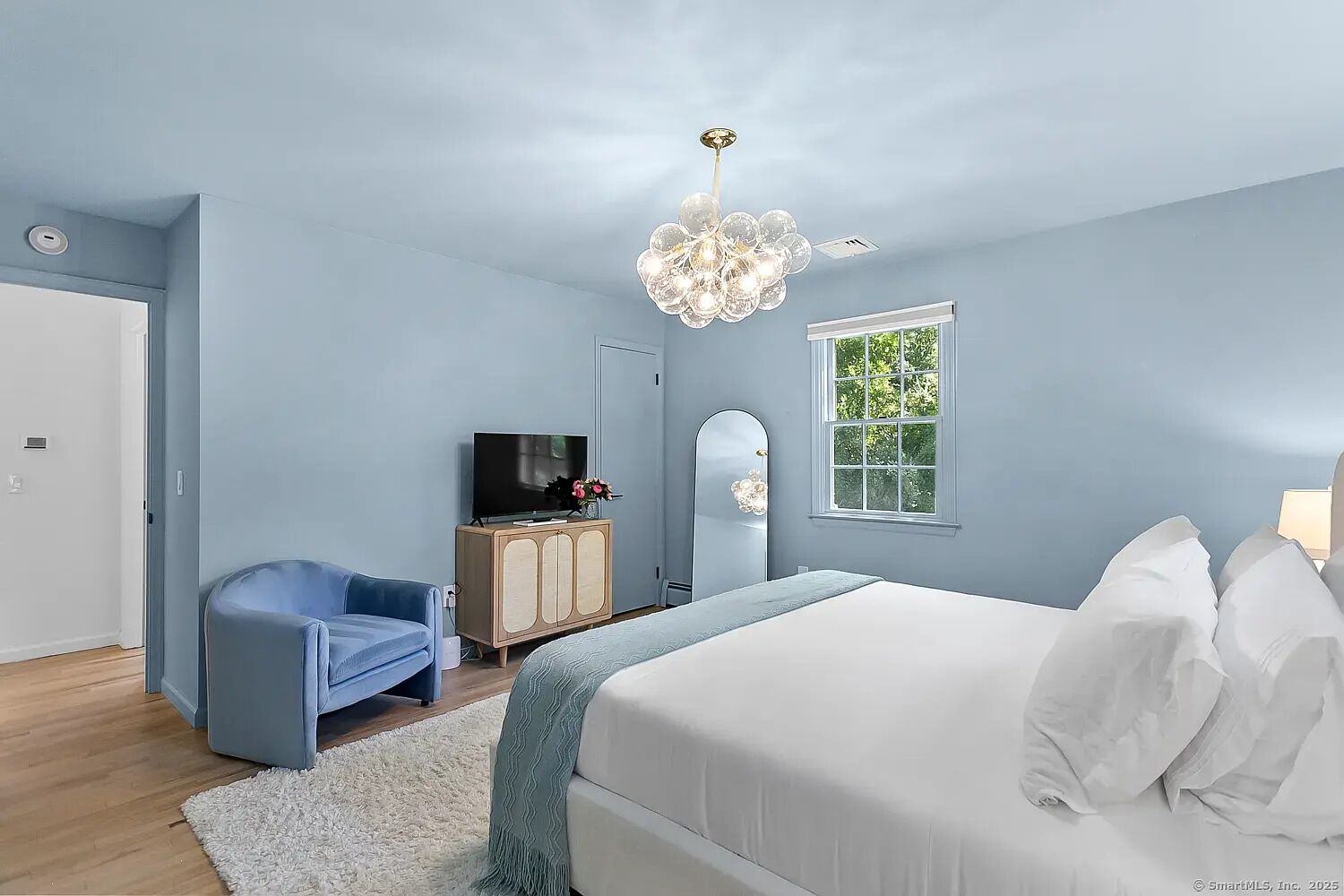 Un dormitorio con una cama grande, un televisor sobre un mueble de madera, una silla azul y una ventana con vista a árboles.