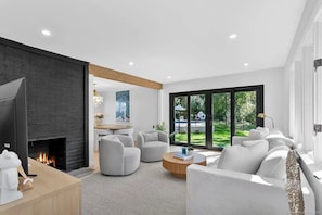 Un salon moderne avec une cheminée, des meubles blancs et une vue sur un jardin grâce à de grandes fenêtres.