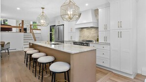 Une cuisine moderne avec des meubles blancs, un îlot central doté de tabourets, des appareils en acier inoxydable et des luminaires suspendus aux formes géométriques.