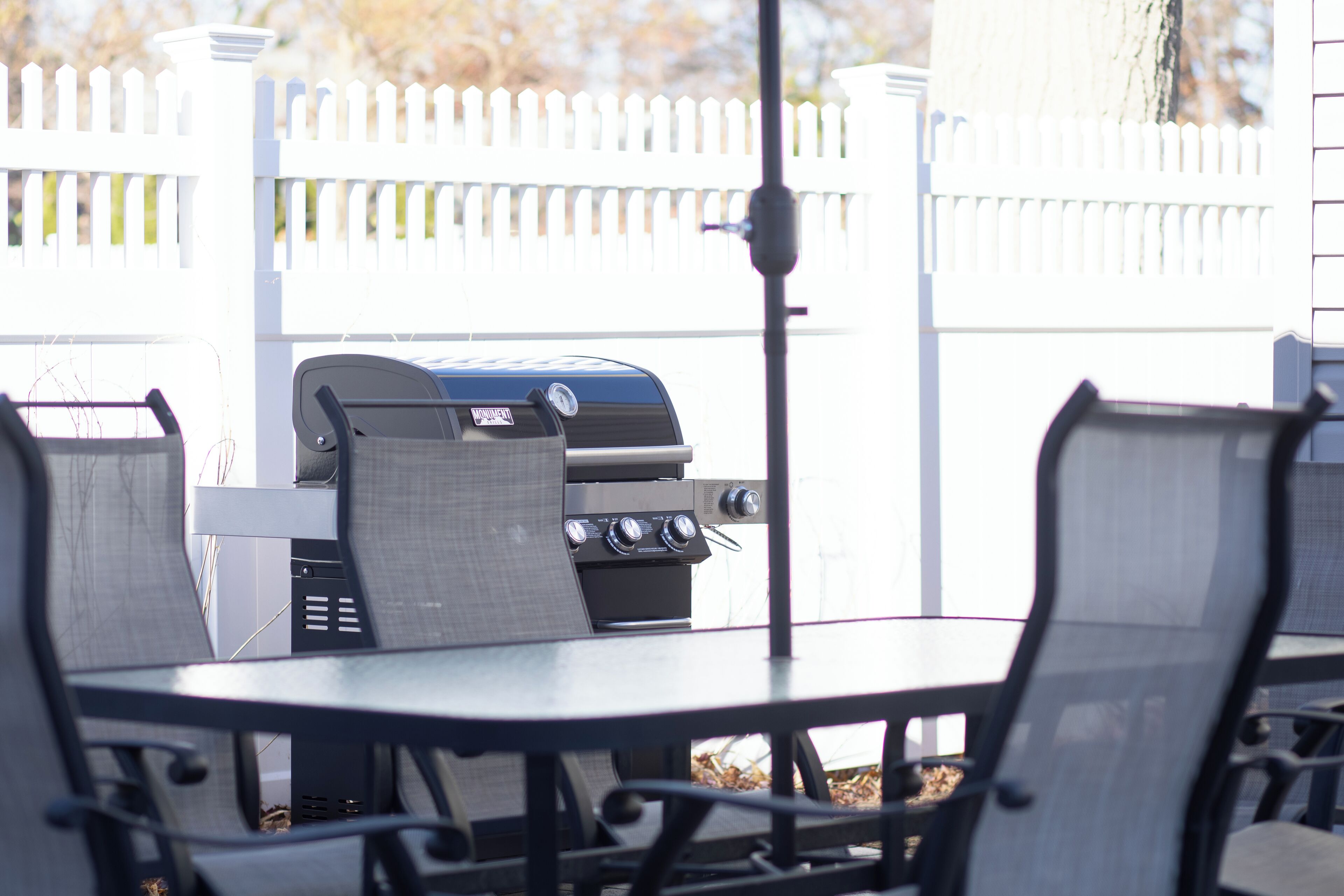 Un patio al aire libre con una parrilla, sillas y un cercado de piquetes blancos.