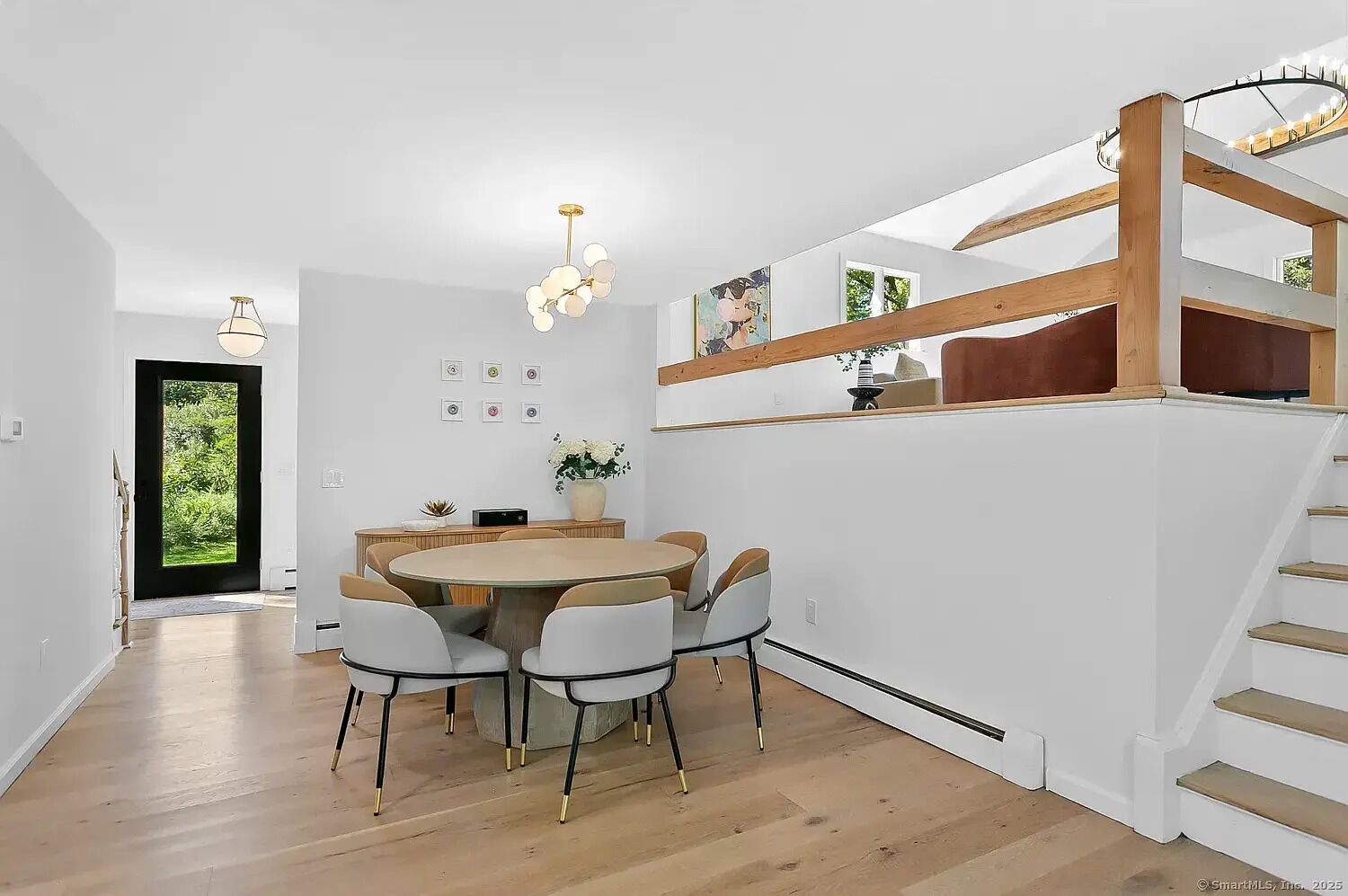 A modern kitchen with a wooden staircase, a dining area with a round table and chairs, and a view of greenery outside.