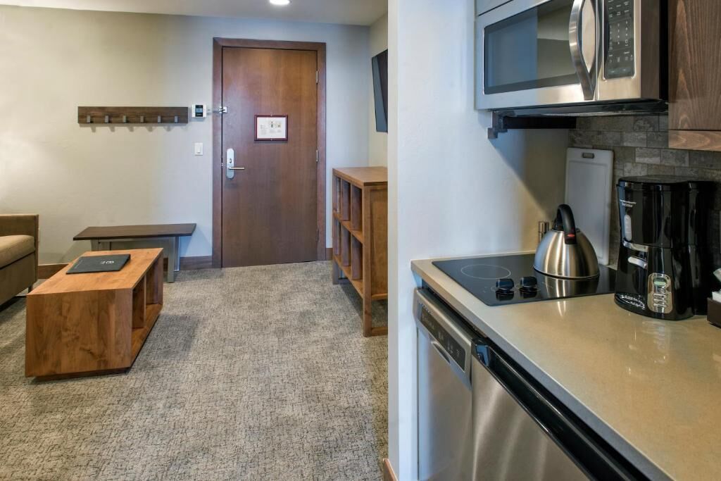 A compact kitchen with a microwave, coffee maker, and dishwasher. There is a wooden bench and a small table in the living area.