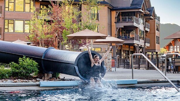 Eine Person saust eine Wasserrutsche in einen Poolbereich, im Hintergrund ist ein Gebäude zu sehen.