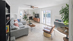 Un salon spacieux avec un canapé gris, une table basse et un téléviseur. On y trouve également une cheminée, un ventilateur de plafond et une grande fenêtre.